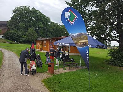 Golf event scene with branded tent and flag by a grassy path, people preparing golf gear, surrounded by lush trees and greenery.