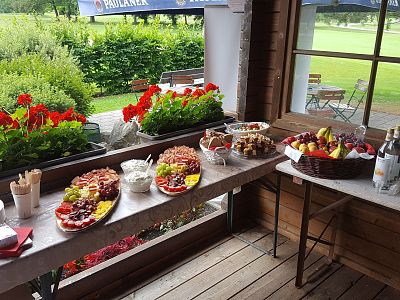A buffet table outdoors with various platters of fruits, snacks, and drinks set against a garden backdrop, next to a wooden building.