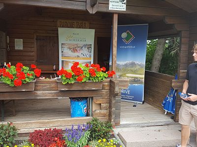 A quaint wooden kiosk with red flowers and colorful banners, evoking a serene, welcoming atmosphere. A person stands nearby, holding an object.