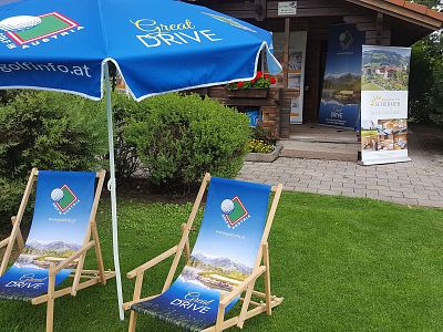 Two wooden deck chairs and a blue umbrella with 'Great Drive' branding on a lush lawn near a charming cottage, surrounded by greenery.