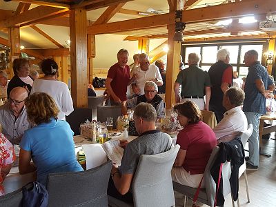 A group of people socializing in a restaurant with wooden decor and large windows, engaged in conversation around tables, creating a warm atmosphere.