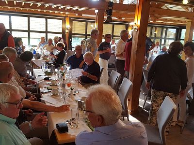 A bustling restaurant scene with numerous people sitting and standing, engaged in conversations at tables. The ambiance suggests a lively social gathering.