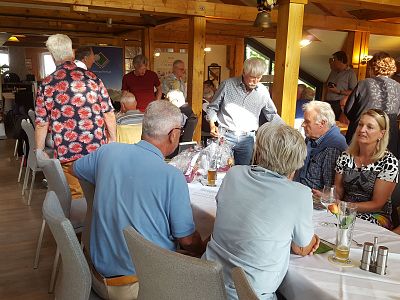 Group of people seated around a table in a restaurant with wooden interiors, engaged in conversation. Plates and glasses are visible, creating a cozy atmosphere.