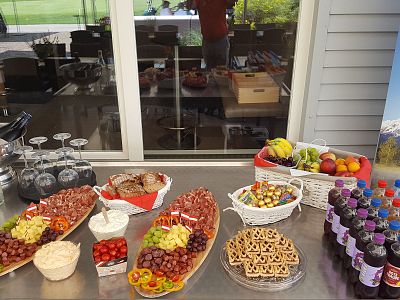 A buffet table with fruits, a basket of snacks, bottled drinks, glasses, and a tray of chocolates. In the background, a window reflects the scene.