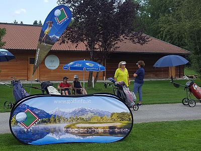 Golf course setup with promo banners, golf carts, and two people talking near a wooden building amidst greenery.