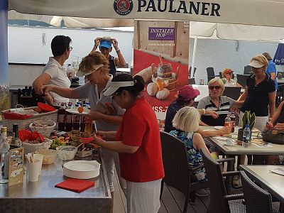 People at an outdoor café under large umbrellas, engaging with food and drinks. The atmosphere is lively and social, with a staff member preparing a table.