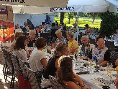 A lively group dining outdoors under umbrellas with food and drinks on a long table, surrounded by greenery and a relaxed ambiance.