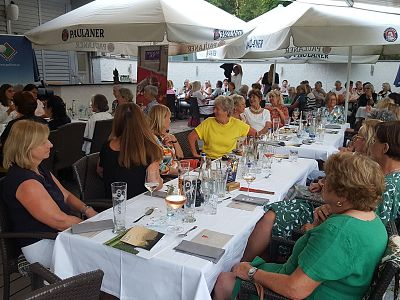 Group of people sitting at outdoor tables under large umbrellas, engaged in conversation, creating a relaxed social gathering atmosphere.