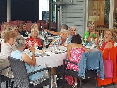 A group of women sitting around a table outdoors, engaged in conversation. Glasses and dishes are on the table, indicating a meal or gathering.