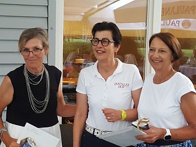 Three women standing together, all smiling. They are holding papers and appear to be at an event or celebration, wearing smart casual attire.