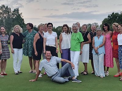 Group of people smiling on a lawn, some standing and one person sitting on the ground. Trees and a cloudy sky are in the background.