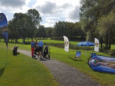 People gather at a golf course with flags and banners on a sunny day. Lush greenery surrounds the area, with a clear path in the foreground.