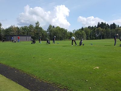 Several people practicing golf on a large grassy area under a partially cloudy sky, surrounded by trees and distant bushes.