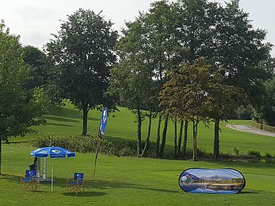 A golf course with lush green grass, trees, and event banners under a partly cloudy sky. Chairs and umbrellas are arranged near colorful banners.
