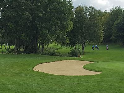 A golf course with a sand bunker, green grass, and tall trees under a partly cloudy sky.