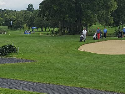 Golfers on a green course with trees, a sand bunker, and golf carts. The landscape provides a serene outdoor setting for the players.