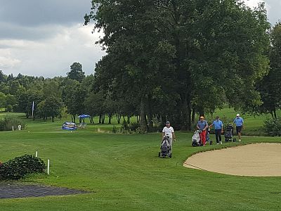Golfers on a green course near a sand bunker, surrounded by trees and under a cloudy sky.