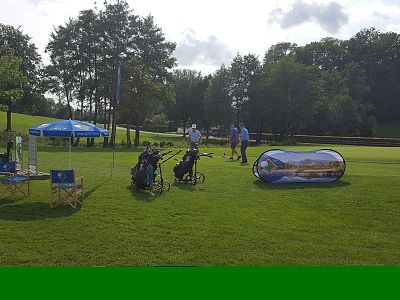 Golf course scene with bags, stands, and people on green grass under a cloudy sky, surrounded by trees.