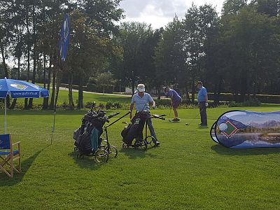 Golfers are seen on a green field with trees, preparing to play. A promotional banner and umbrella suggest a professional event atmosphere.