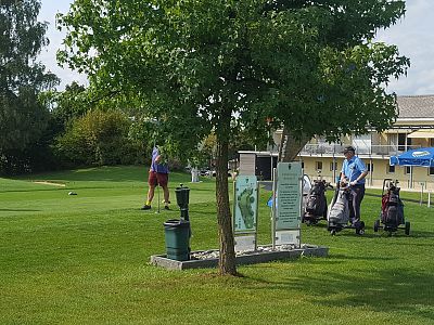 Two golfers on a green course with trees and a building. Bright day with well-maintained grass, golf equipment visible, and a relaxing atmosphere.