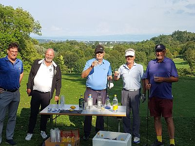 Five men on a golf course, smiling with drinks, standing by a table with bottles. Greenery and a scenic landscape spread out behind them.