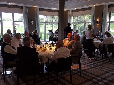 People seated at round tables in an elegantly decorated room with large windows, engaging in conversation and dining formally. Natural light illuminates the scene.