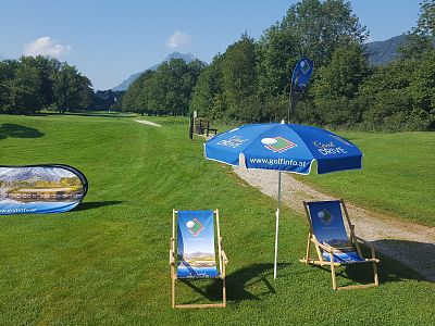 Golf course with two deck chairs and an umbrella on a grassy area, surrounded by trees and hills under a clear blue sky.