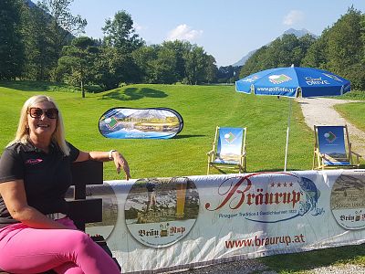 A woman in sunglasses sits on a bench next to a banner with a scenic golf course in the background, green grass, clear sky, and mountain view.
