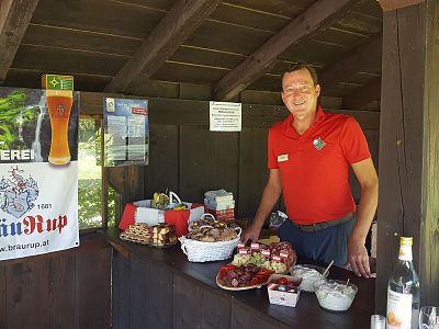 Vendor stands behind a counter with an array of food items in an outdoor market setting. A "BrauRup" poster is visible on the left.