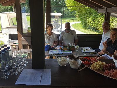 Four people sit at an outdoor wooden table under a roof, with drinks and snacks, surrounded by greenery. A golf cart is seen in the background.