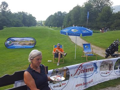 A person relaxes in a sun chair with a blue umbrella on a golf course. Nearby, a woman sits with plates. Brewery signs and golf equipment are visible.