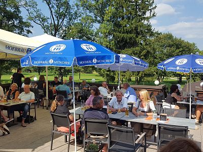 People at outdoor restaurant seated under blue "Hofbräu München" umbrellas, enjoying meals and drinks on a sunny day. Trees and grass in the background.
