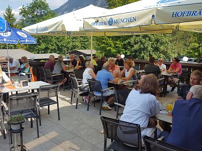 A crowded outdoor beer garden with people seated under umbrellas, enjoying drinks. Trees in the background add greenery to the sunny setting.