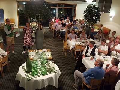 A diverse group of people seated at tables in a restaurant, listening to a speaker in traditional attire. The decor is cozy with plants and soft lighting.