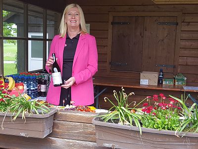 Person in pink blazer at a wooden market stall with flowers and bottled drinks.