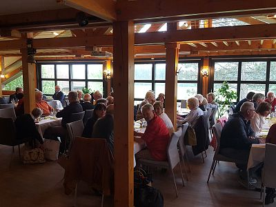 A spacious hall with wooden beams where people of various ages are seated at tables, engaged in conversations, with large windows letting in natural light.
