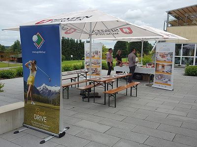 Outdoor event setup with tables, benches, umbrellas, and banners showing a golfer. Background includes a patio space, trees, and a building.