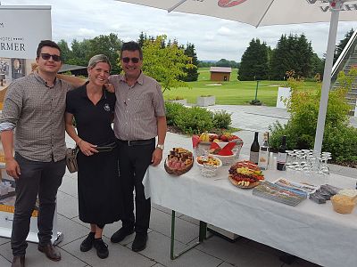 Three people stand by a table with snacks and drinks, under umbrellas, on a patio overlooking green grass and trees.