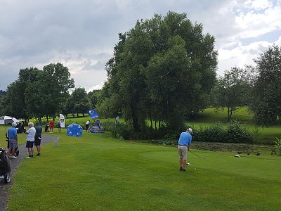People in a park playing golf on a bright day with green grass and trees around. A golfer in the foreground is taking a shot.