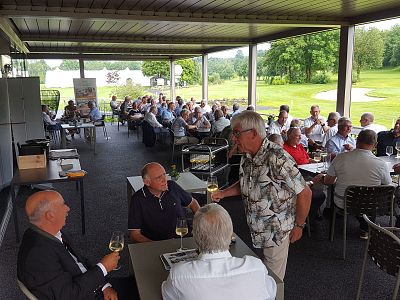 People seated at tables in a covered patio, enjoying drinks and conversation. A lush golf course landscape is visible in the background.
