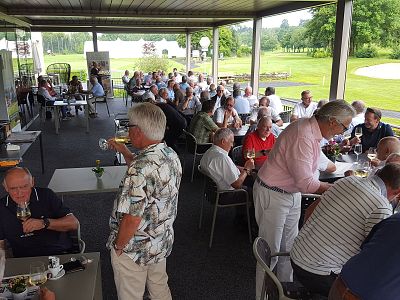 People gathered on a covered patio with tables, chatting and enjoying food and drinks, surrounded by green lawns and trees.