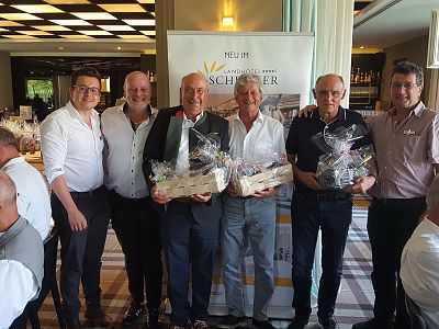 A group of men in a formal setting holding gift baskets, standing in front of a banner. They are smiling and dressed casually, with a restaurant background.