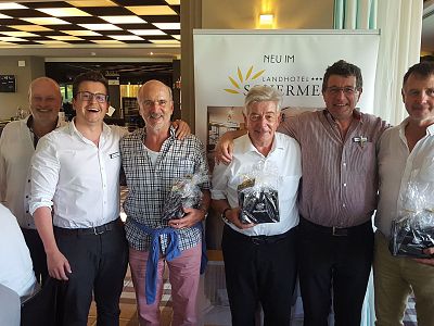Six men standing side by side, smiling at the camera at a celebratory event, with some holding gift bags.