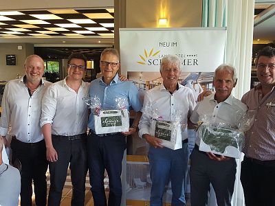 Six men stand smiling, three holding gift baskets in front of a golf resort banner. They appear to be at an event or celebration in a well-lit room.