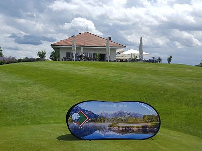 A lush golf course with a scenic promotional sign in the foreground. A clubhouse with a patio and umbrellas is in the background under a partly cloudy sky.