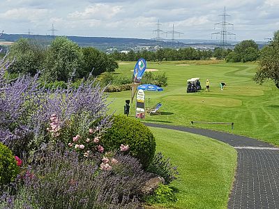 A scenic golf course with manicured grass, blooming flowers, and players in the distance. Hills and power lines are visible under a partly cloudy sky.