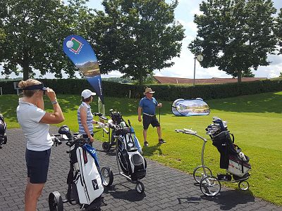 Golfers with bags and trolleys ready to play on a sunny course. Trees and promotional flags are in view.