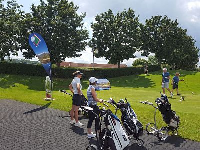 Golfers in sport attire stand on a grassy course with golf bags. The scene is sunny with trees and a blue sky in the background.