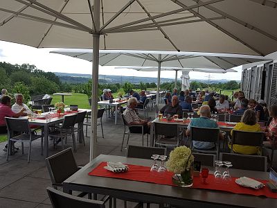 A busy outdoor restaurant with diners sitting at tables under umbrellas, enjoying their meals. The scene overlooks a green landscape and the sky is partly cloudy.
