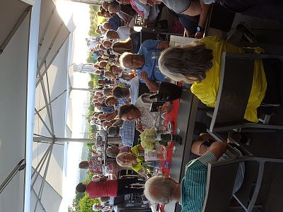 People seated at tables under umbrellas, engaging in lively conversation and dining outdoors in a sunny environment.